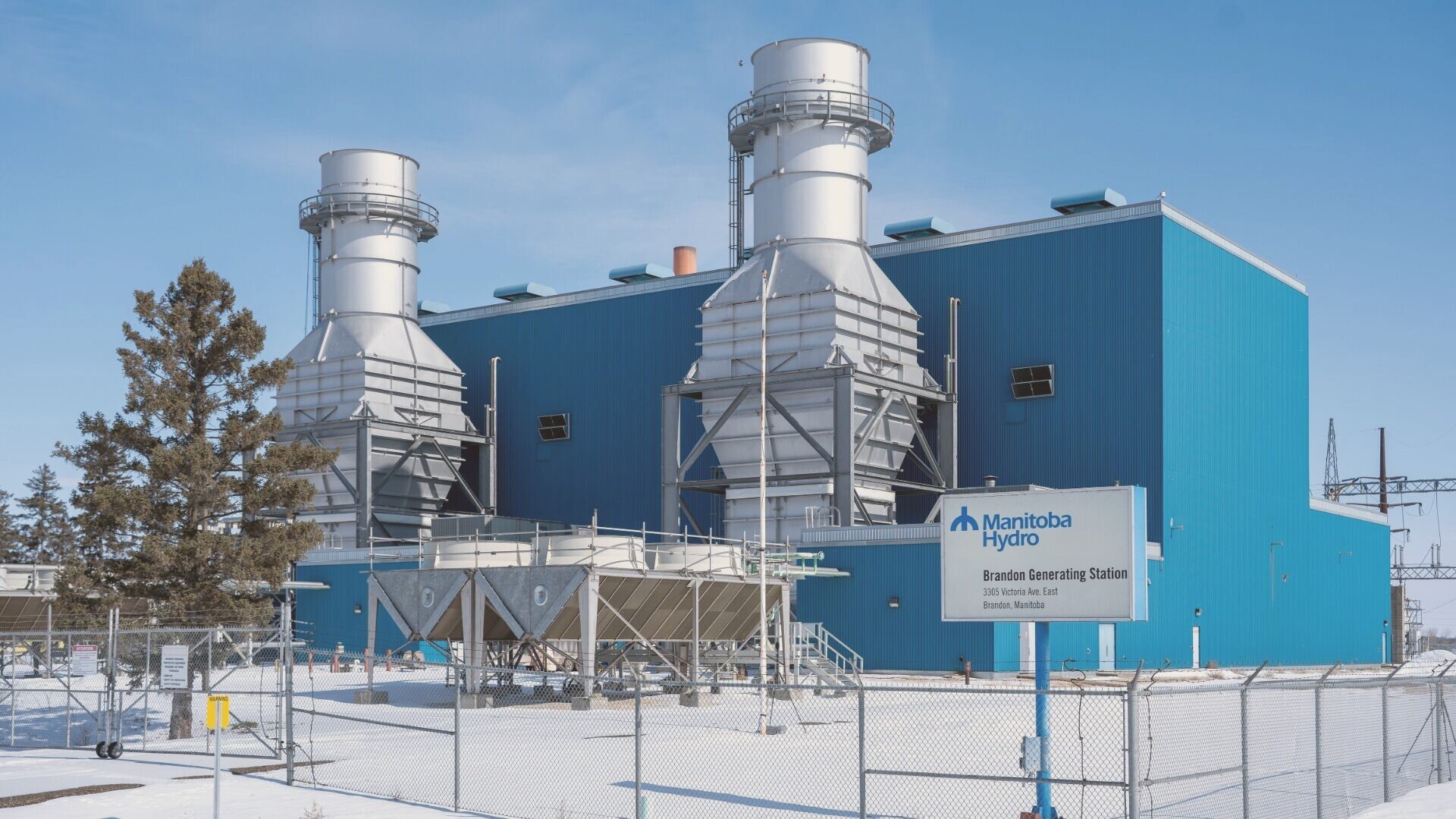 The Brandon Generating Station: a blue industrial building with two silver gas turbine exhaust stacks behind a chain-link fence in snow, with a Manitoba Hydro sign in front