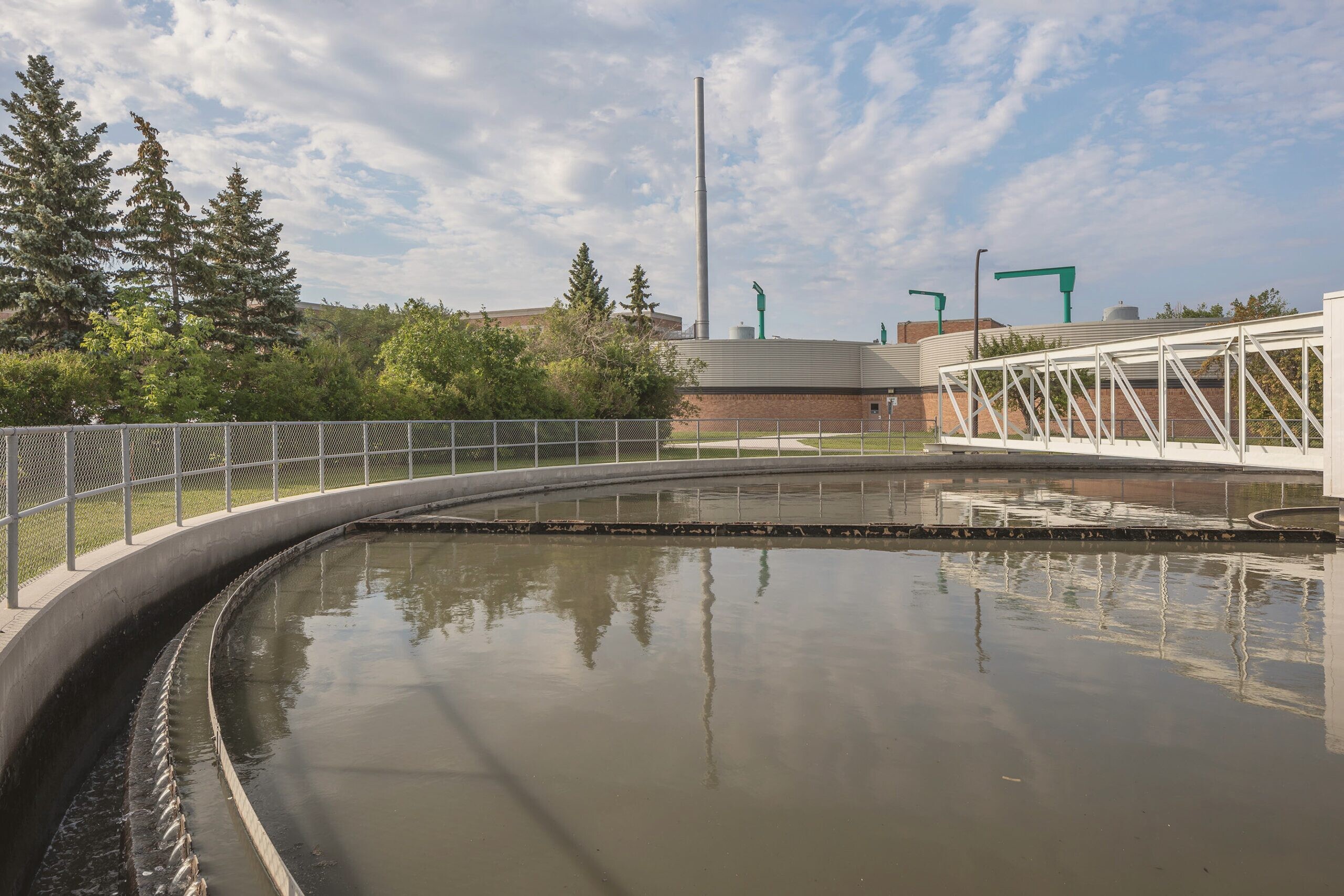 A circular clarifier pool of warm treated water at the North End Water Pollution Control Centre, with the plant buildings and stack behind